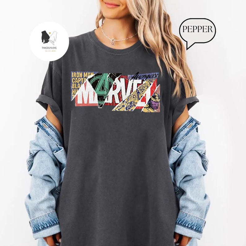 Op de afbeelding: Donkergrijs T-shirt met het Marvel-logo in een kleurrijk, geometrisch ontwerp. Het ontwerp bevat tekstelementen zoals "Iron Man", "Captain" en "Avengers". Het shirt heeft opgerolde mouwen en wordt gedragen met een spijkerjasje.