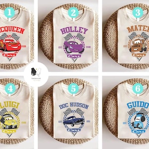May include: Six white t-shirts featuring different Cars characters. Each shirt has a chequered flag design and the text "Cars" below the character. The characters are McQueen, Holley, Mater, Luigi, Doc Hudson, and Guido.
