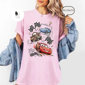 May include: Pink t-shirt featuring a colorful graphic of animated race cars, checkered flags, and racing elements. The design includes a red race car, a blue car, and a brown tow truck. The shirt has a crew neck and short sleeves.