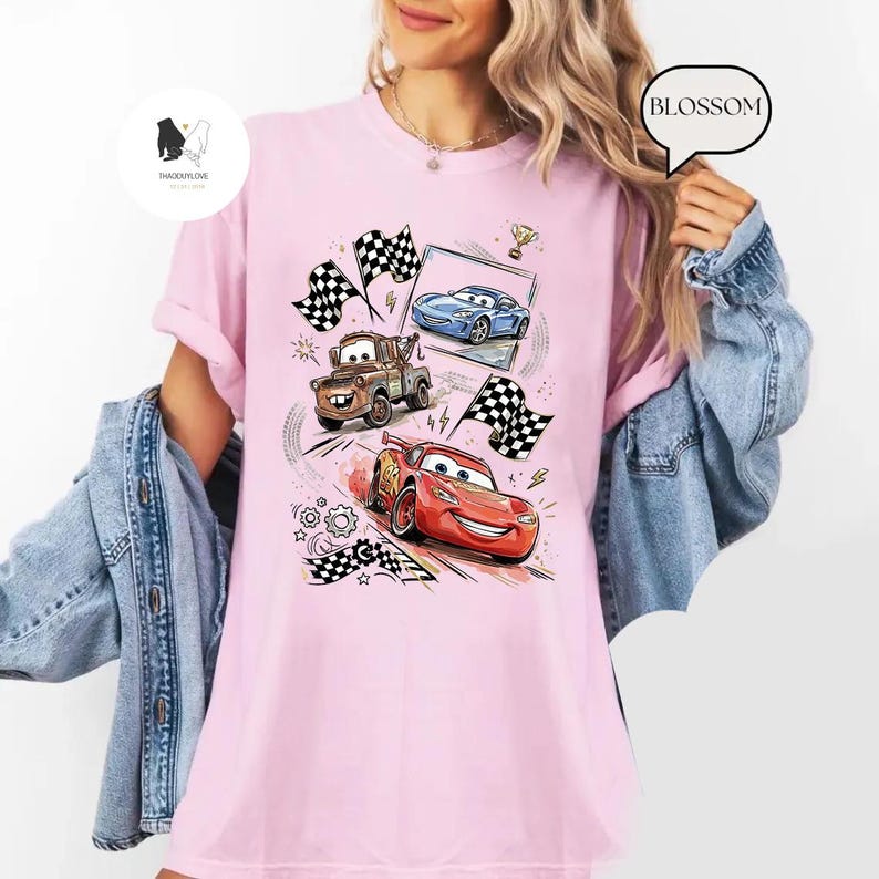 May include: Pink t-shirt featuring a colorful graphic of animated race cars, checkered flags, and racing elements. The design includes a red race car, a blue car, and a brown tow truck. The shirt has a crew neck and short sleeves.