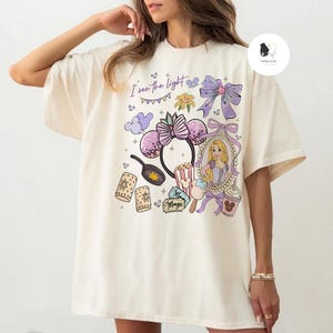 May include: Cream-colored t-shirt featuring a colorful graphic design. The design includes a portrait of a princess, Minnie Mouse ears, popcorn, and the text "I see the light". The shirt has short sleeves and a relaxed fit.