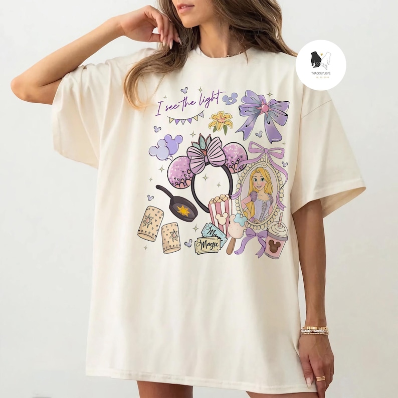 May include: Cream-colored t-shirt featuring a colorful graphic design. The design includes a portrait of a princess, Minnie Mouse ears, popcorn, and the text "I see the light". The shirt has short sleeves and a relaxed fit.