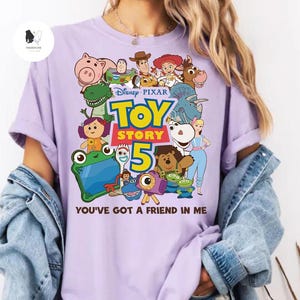 May include: A lilac t-shirt with a vibrant graphic of characters from the Disney Pixar film Toy Story 5. The design includes the film's title and the words "You've Got a Friend in Me".