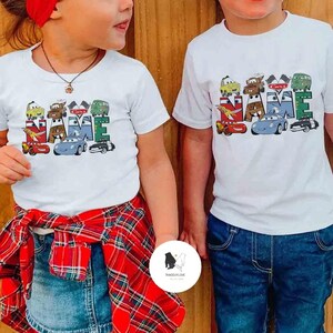 K&ouml;nnte beinhalten: Zwei wei&szlig;e T-Shirts mit einem farbenfrohen Autodesign mit der Aufschrift "NAME". Das Design umfasst verschiedene Cartoon-Autos in Rot, Blau, Gr&uuml;n und Braun. Die Shirts werden von Kindern getragen.