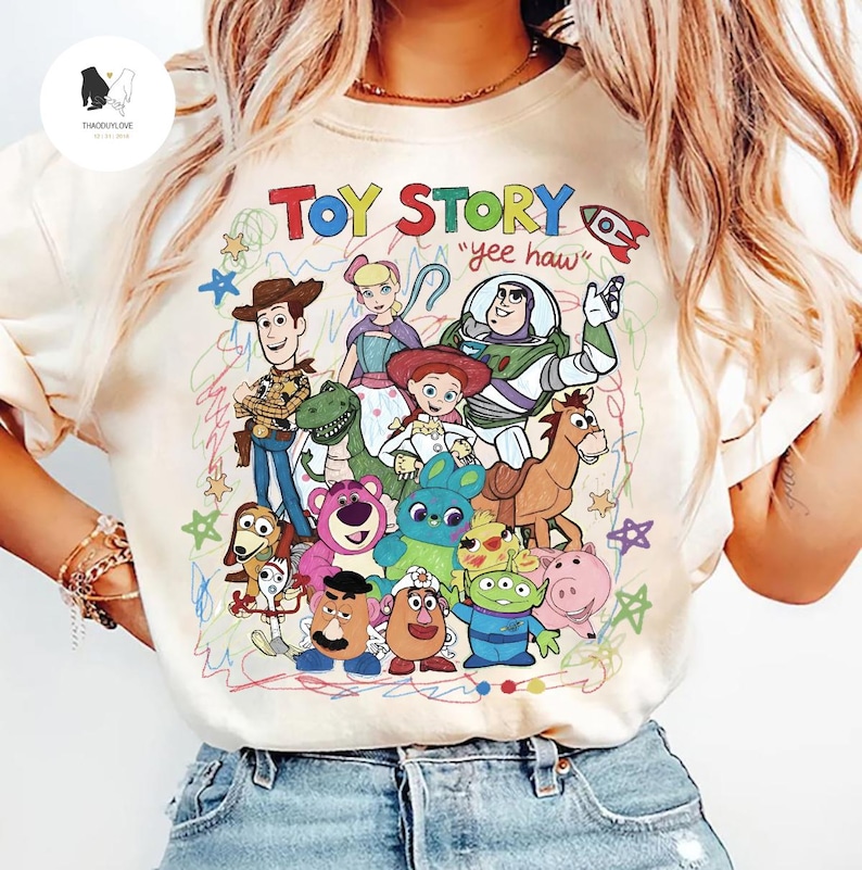 May include: A cream-colored t-shirt featuring a colorful graphic print of Toy Story characters including Woody, Buzz Lightyear, Jessie, and others. The text "TOY STORY" appears at the top in multi-colored letters, with "yee haw" written below.