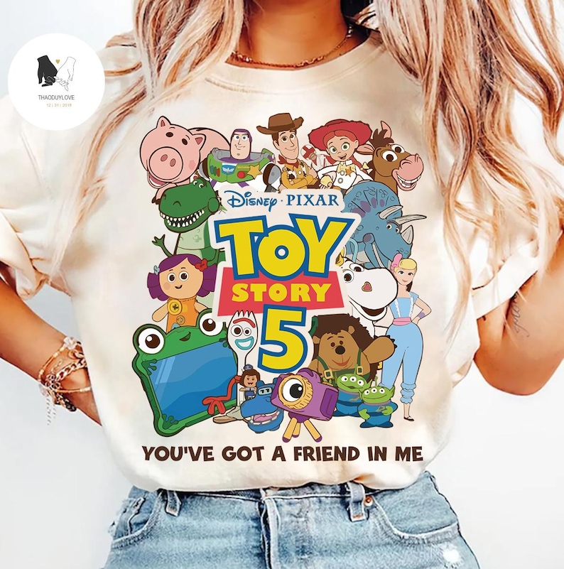 May include: Cream-coloured t-shirt with a vibrant graphic of characters from Disney Pixar's Toy Story 5. The design showcases Woody, Buzz Lightyear, and other popular characters, along with the phrase "YOU'VE GOT A FRIEND IN ME".