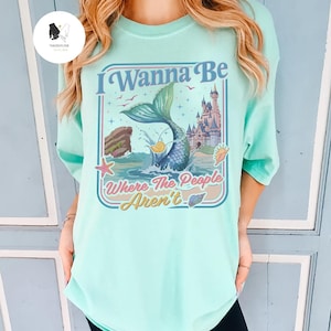 May include: A light teal t-shirt with a colorful graphic featuring a mermaid tail, a castle, and the text "I Wanna Be Where The People Aren't". The design includes ocean elements and a retro style.