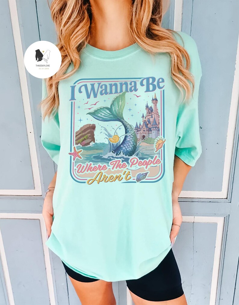 May include: A light teal t-shirt with a colorful graphic featuring a mermaid tail, a castle, and the text "I Wanna Be Where The People Aren't". The design includes ocean elements and a retro style.
