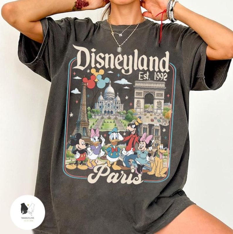 May include: Dark gray t-shirt featuring a colorful graphic of Disneyland Paris. The design includes Mickey Mouse, Minnie Mouse, Donald Duck, and Goofy, along with iconic landmarks like the Eiffel Tower and Arc de Triomphe. The text "Disneyland Paris Est. 1992" is also visible.