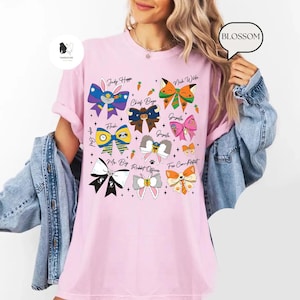 Puede incluir: Una camiseta rosa claro con dise&ntilde;os de lazos coloridos y nombres de personajes como "Judy Hoppe" y "Nick Wilde". Los lazos tienen varios patrones y colores, incluyendo azul, verde y naranja. La palabra "BLOSSOM" est&aacute; en una burbuja.