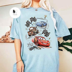 May include: Light blue t-shirt featuring a colorful graphic of animated race cars, including a red car, a blue car, and a brown tow truck. The design includes checkered flags, a trophy, and lightning bolts.