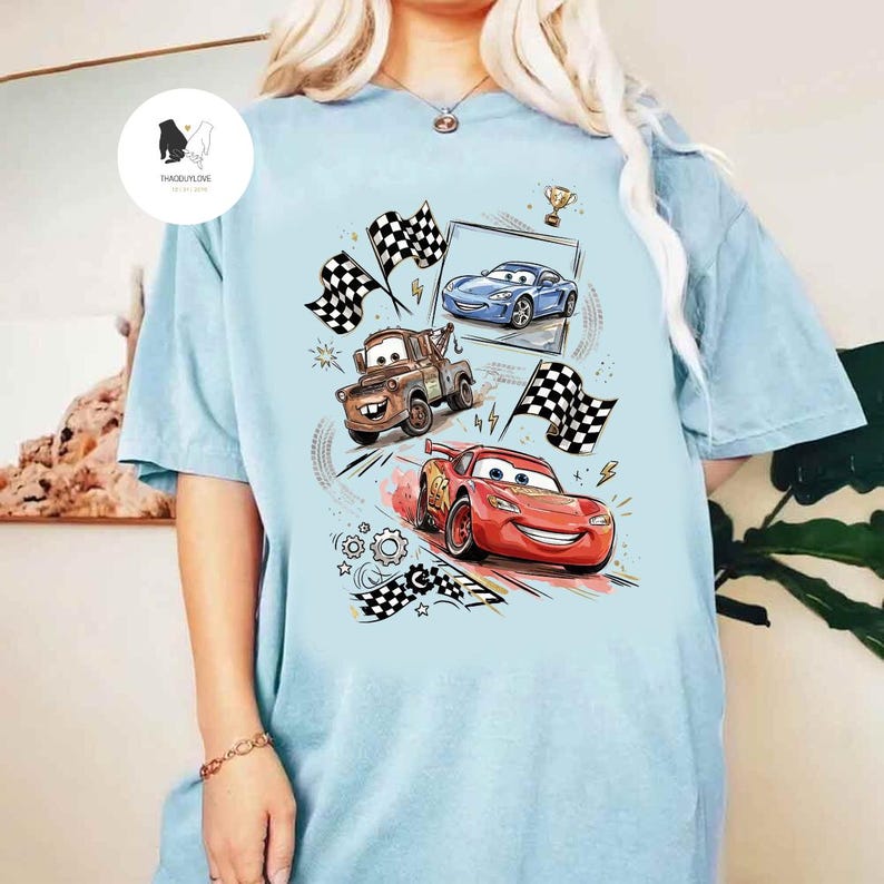 May include: Light blue t-shirt featuring a colorful graphic of animated race cars, including a red car, a blue car, and a brown tow truck. The design includes checkered flags, a trophy, and lightning bolts.