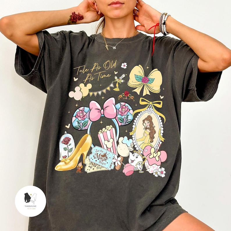 May include: Dark gray t-shirt featuring a colorful graphic design with the text "Tale As Old As Time." The design includes a pink bow, a portrait of a princess, and other themed elements. The shirt has short sleeves and a relaxed fit.