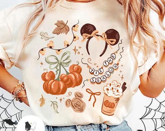 Könnte beinhalten: Cremefarbenes T-Shirt mit herbstlichen Grafiken. Mit Kürbissen, Mickey Mouse Ohren, Blättern und einer Kaffeetasse. Der Text lautet "Tis the Season". Ein festliches Design für den Herbst.