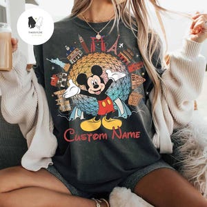May include: Dark gray t-shirt featuring Mickey Mouse with arms outstretched in front of a colorful globe design. The shirt includes the text "Custom Name" in red, and iconic landmarks. A fun, personalized Disney-themed apparel.