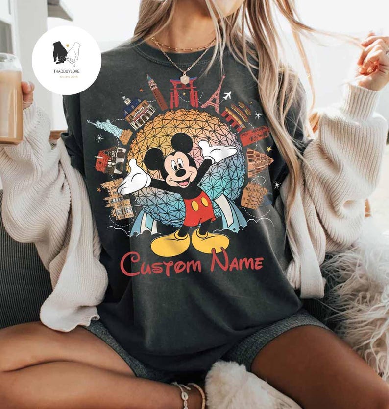 May include: Dark gray t-shirt featuring Mickey Mouse with arms outstretched in front of a colorful globe design. The shirt includes the text "Custom Name" in red, and iconic landmarks. A fun, personalized Disney-themed apparel.