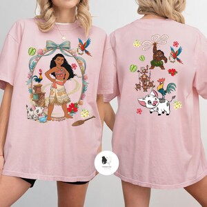 以下が含まれることがあります： 明るいピンク色のTシャツ。前面には、漫画のキャラクター、オウム、豚、鶏が装飾的なフレームの中に描かれたカラフルなグラフィックデザイン。背面にも同様のデザインが施されています。