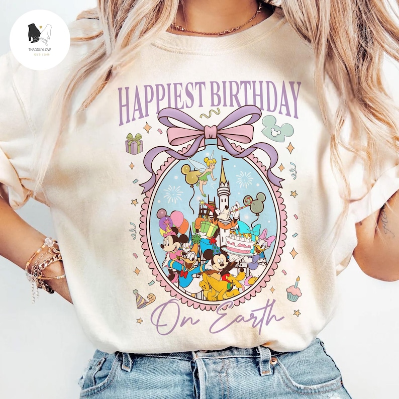 May include: Cream-colored t-shirt featuring a colorful graphic with Disney characters, a castle, and the text "Happiest Birthday On Earth." The design includes Mickey Mouse, Minnie Mouse, Donald Duck, and other characters. The graphic is framed by a light purple bow and decorative elements.