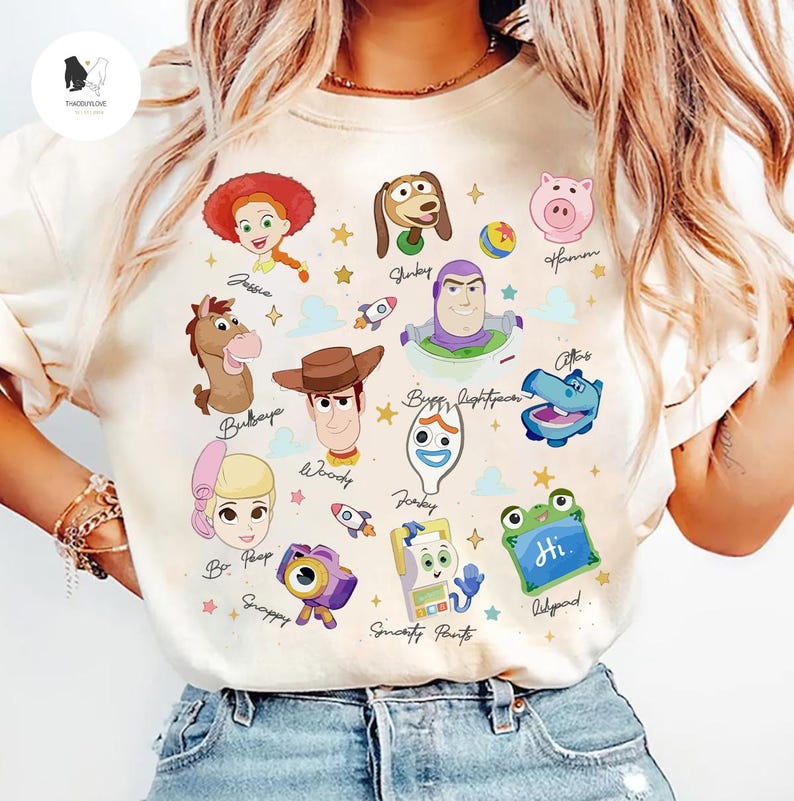 May include: Cream-colored t-shirt featuring colorful cartoon illustrations of characters from the movie Toy Story. The design includes images of Woody, Buzz Lightyear, Jessie, and others, with their names written below.