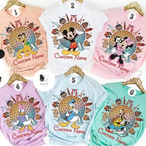 May include: Six colorful t-shirts featuring Disney characters and a globe design with landmarks. Each shirt has a different character: Pluto, Mickey Mouse, Minnie Mouse, Daisy Duck, Donald Duck, and Goofy. The text "Custom Name" is printed on each shirt.