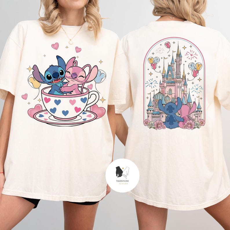 May include: Cream-colored t-shirt featuring a cartoon illustration of Stitch and Angel in a teacup with hearts. The back of the shirt shows a castle scene with Stitch and Angel. The shirt has a round neck and short sleeves.