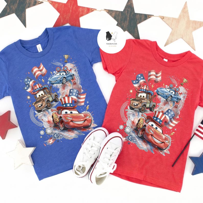 Op de afbeelding: Een blauw t-shirt en een rood t-shirt, elk met een grafische print van cartoonraceauto's met patriottische hoge hoeden, vergezeld door Amerikaanse vlaggen, een gouden trofee en abstracte kleurspatten. Een paar witte sneakers ligt ertussen.