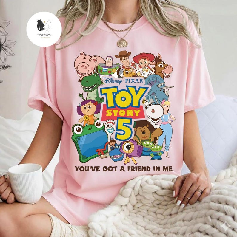 May include: Pale pink t-shirt with a vibrant Toy Story graphic, showcasing characters like Woody and Buzz Lightyear. The design includes "TOY STORY 5" and the phrase "YOU'VE GOT A FRIEND IN ME". A fun, colourful design.