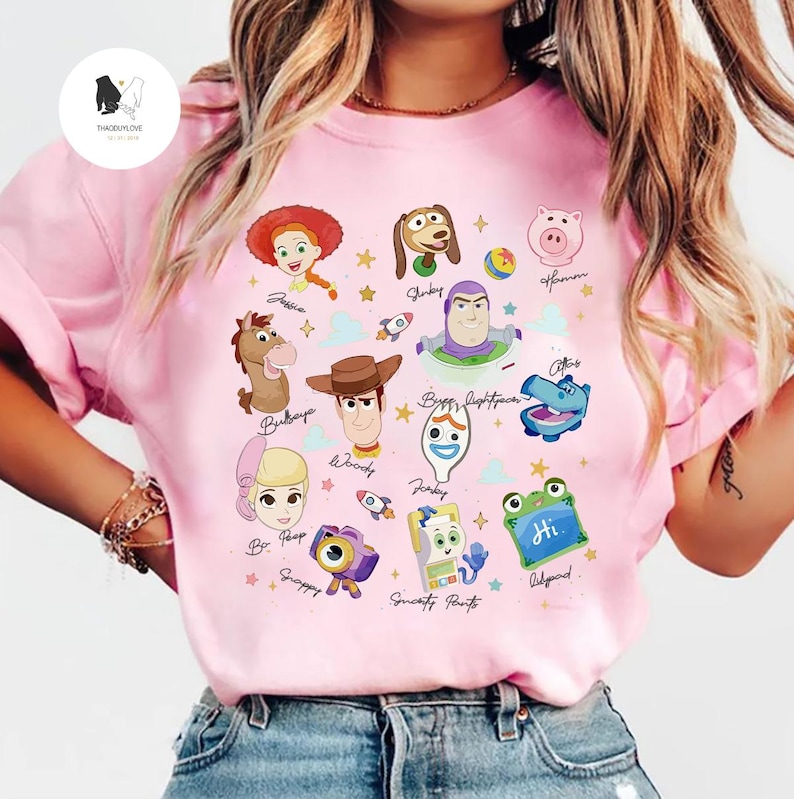 May include: Pink t-shirt featuring colorful illustrations of characters from the animated movie Toy Story. The design includes characters like Woody, Buzz Lightyear, and Jessie, with their names written below. The shirt has a crew neck and short sleeves.