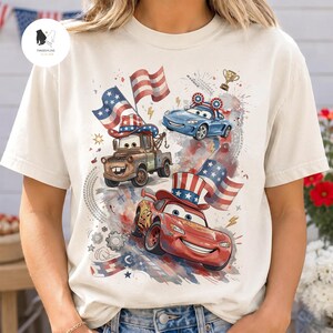 Op de afbeelding: Een cr&egrave;mekleurig t-shirt met een grafische print van drie geanimeerde auto's met Amerikaanse vlagmotieven. Het ontwerp bevat een bruine takelwagen met een vlaghoed, een rode raceauto met een vlag-hoge hoed en een blauwe auto met vlag-oren.