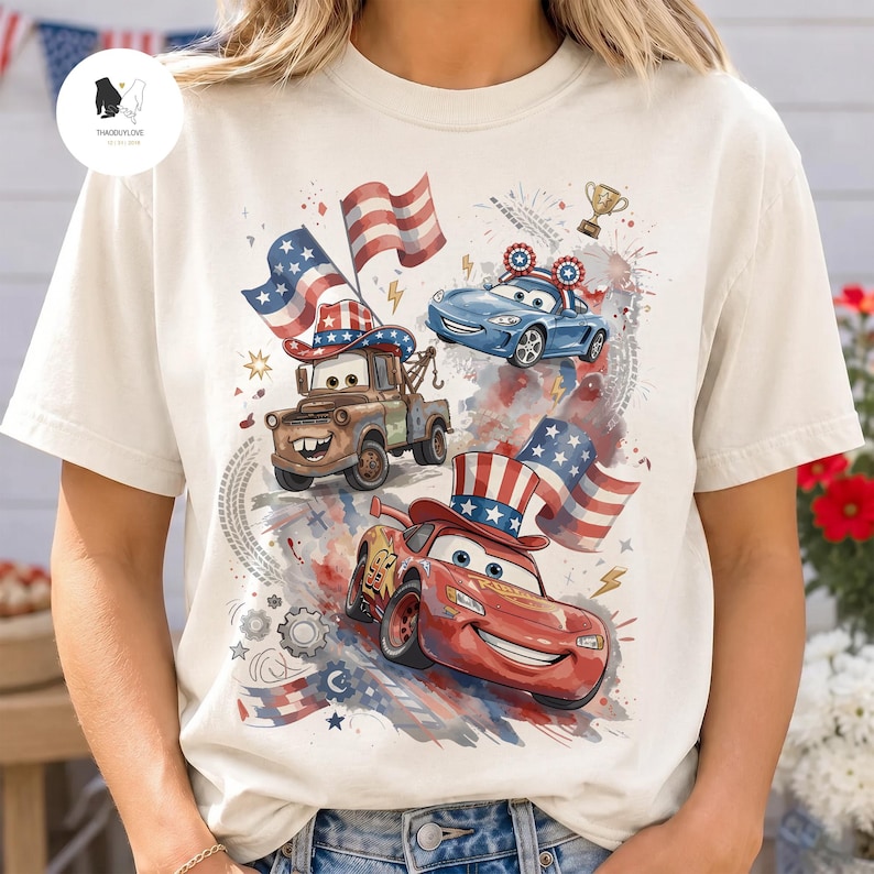 Op de afbeelding: Een cr&egrave;mekleurig t-shirt met een grafische print van drie geanimeerde auto's met Amerikaanse vlagmotieven. Het ontwerp bevat een bruine takelwagen met een vlaghoed, een rode raceauto met een vlag-hoge hoed en een blauwe auto met vlag-oren.