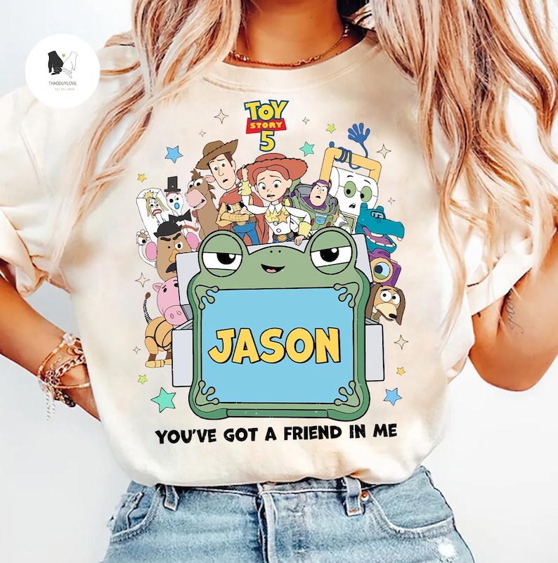 May include: Cream-colored t-shirt featuring a colorful graphic of characters from Toy Story 5. The design includes a frog holding a sign that says "JASON" and the phrase "YOU'VE GOT A FRIEND IN ME".