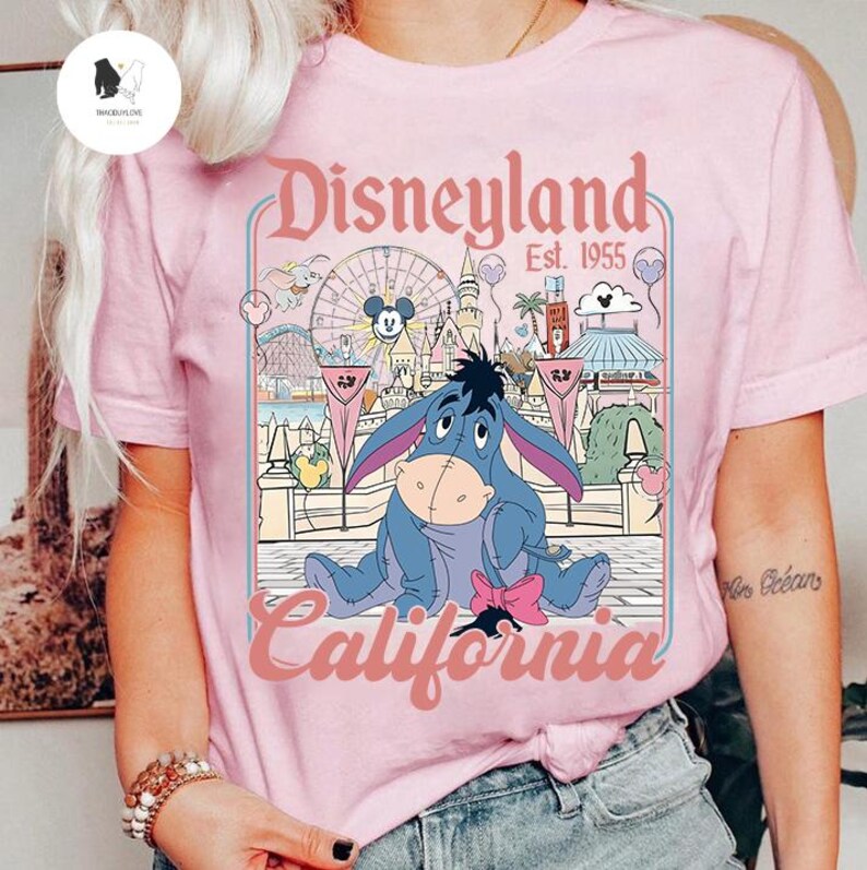 Peut inclure: T-shirt rose clair avec un motif Disneyland. Le motif comprend Eeyore, l'&acirc;ne, et le texte "Disneyland California Est. 1955". Le motif pr&eacute;sente une grande roue et d'autres attractions du parc.