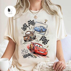 May include: Cream-colored t-shirt featuring a colorful graphic of animated race cars, including a red car, a blue car, and a brown tow truck. The design includes checkered flags, a trophy, and racing details.