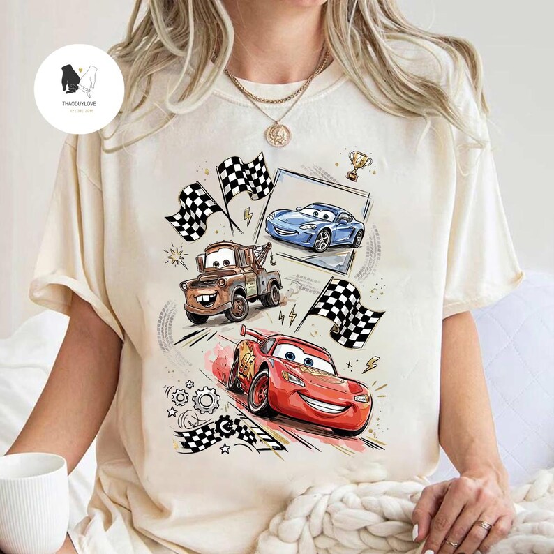 May include: Cream-colored t-shirt featuring a colorful graphic of animated race cars, including a red car, a blue car, and a brown tow truck. The design includes checkered flags, a trophy, and racing details.
