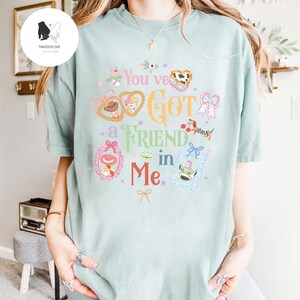 May include: A light green t-shirt featuring the phrase "You've Got a Friend in Me" in a whimsical font. The design includes cartoon character illustrations in frames and decorative elements in pink, yellow, and blue.