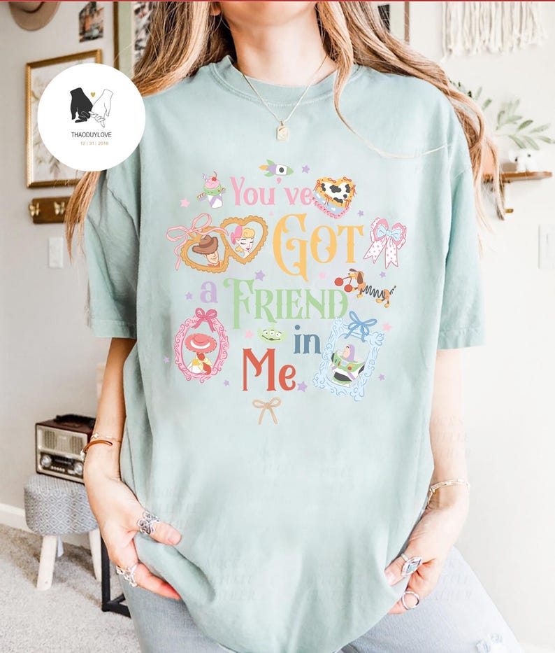 May include: A light green t-shirt featuring the phrase "You've Got a Friend in Me" in a whimsical font. The design includes cartoon character illustrations in frames and decorative elements in pink, yellow, and blue.