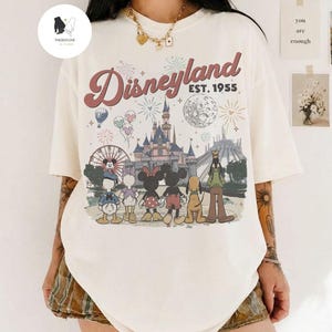 May include: An off-white short-sleeve t-shirt featuring a graphic print of the Disneyland castle, fireworks, and Disney characters including Mickey, Minnie, Donald, Daisy, Pluto, and Goofy. The text reads Disneyland EST. 1955 in a retro font.