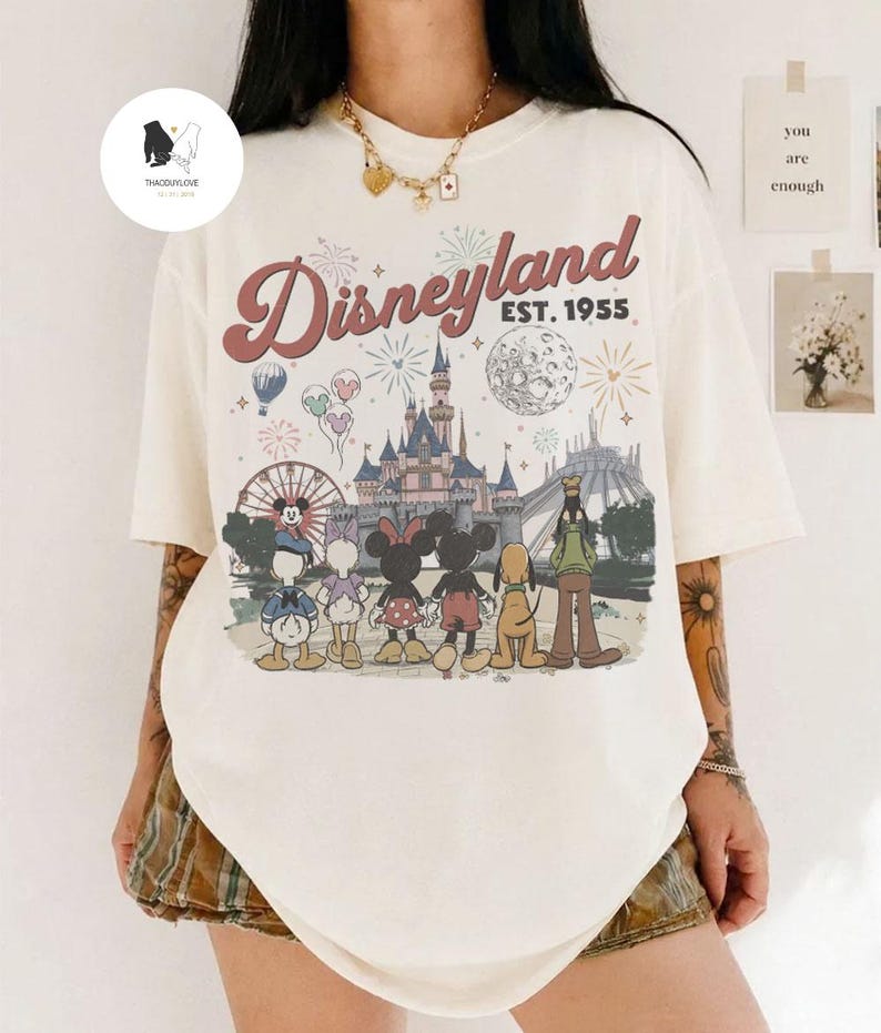 May include: An off-white short-sleeve t-shirt featuring a graphic print of the Disneyland castle, fireworks, and Disney characters including Mickey, Minnie, Donald, Daisy, Pluto, and Goofy. The text reads Disneyland EST. 1955 in a retro font.