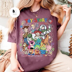 May include: A maroon short-sleeve t-shirt featuring a colorful graphic print of Toy Story characters including Woody, Buzz Lightyear, Jessie, Rex, and others. The design includes the text TOY STORY in multi-colored letters and scribbled line art.