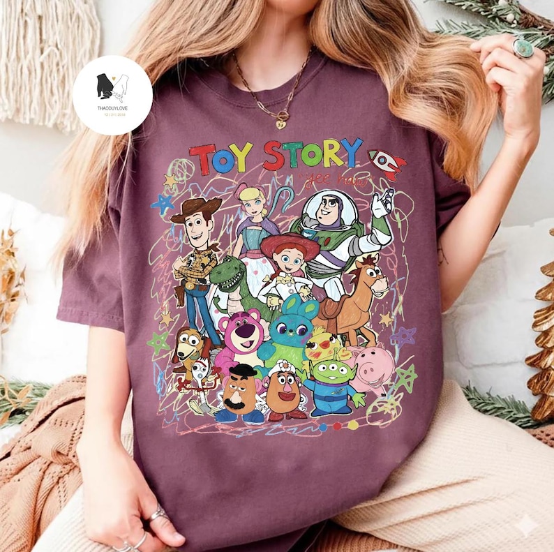 May include: A maroon short-sleeve t-shirt featuring a colorful graphic print of Toy Story characters including Woody, Buzz Lightyear, Jessie, Rex, and others. The design includes the text TOY STORY in multi-colored letters and scribbled line art.
