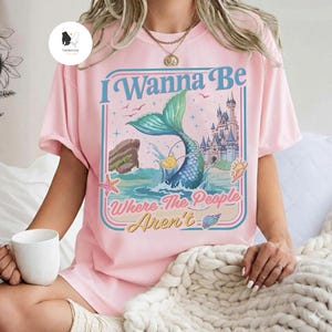 May include: Pink t-shirt with a mermaid graphic and the text "I Wanna Be Where The People Aren't". The graphic features a mermaid tail, ocean waves, a castle, and seashells. The shirt has short sleeves.
