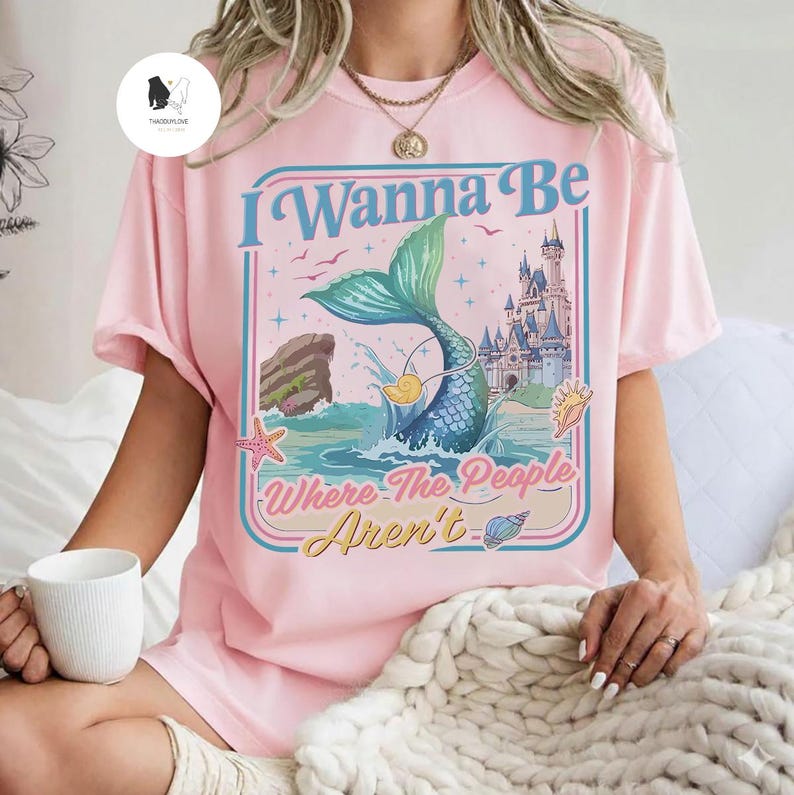 May include: Pink t-shirt with a mermaid graphic and the text "I Wanna Be Where The People Aren't". The graphic features a mermaid tail, ocean waves, a castle, and seashells. The shirt has short sleeves.