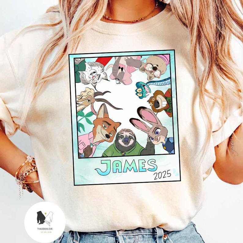 以下が含まれることがあります： ズートピアのキャラクターの漫画のイメージがプリントされたクリーム色のTシャツ。 画像はポラロイドのようにフレームされており、「JAMES」という名前と「2025」という年が水色のフォントで表示されています。 Tシャツは丸いネックラインと半袖です。