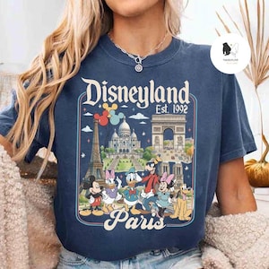 May include: Navy blue t-shirt featuring a Disneyland Paris graphic with Mickey Mouse, Minnie Mouse, Donald Duck, and Goofy. The graphic includes the Eiffel Tower, Arc de Triomphe, and Sacr&eacute;-C&oelig;ur. The text reads "Disneyland Est. 1992 Paris."