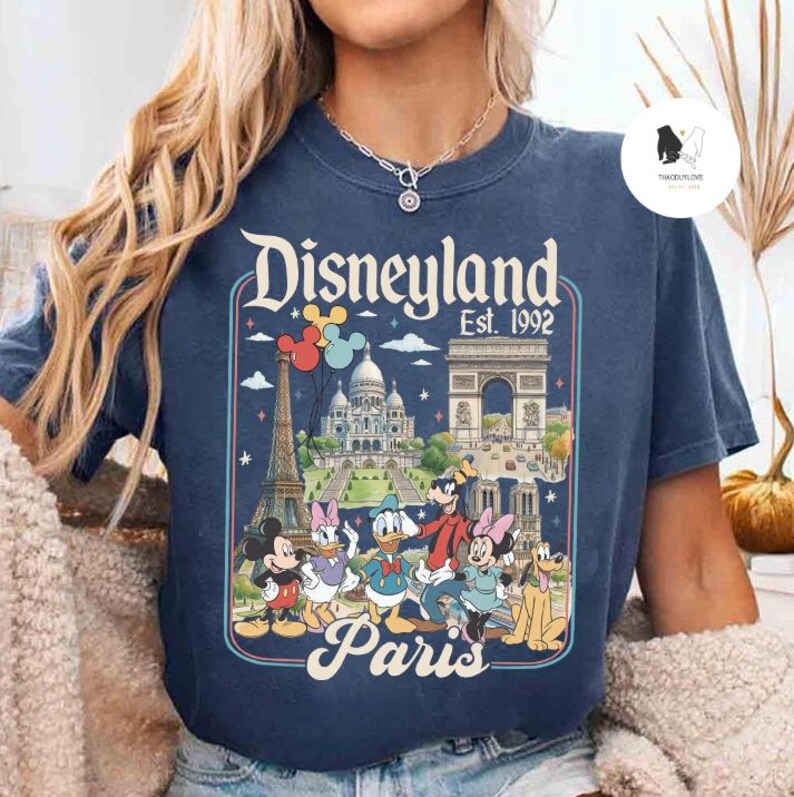 May include: Navy blue t-shirt featuring a Disneyland Paris graphic with Mickey Mouse, Minnie Mouse, Donald Duck, and Goofy. The graphic includes the Eiffel Tower, Arc de Triomphe, and Sacr&eacute;-C&oelig;ur. The text reads "Disneyland Est. 1992 Paris."