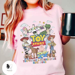 May include: Pink t-shirt with a graphic of the Toy Story characters, including Woody, Buzz Lightyear, Jessie, and Rex. The text "Toy Story" is printed in large letters, and the phrase "You've got a friend in me Est. 1995" is printed below the characters.