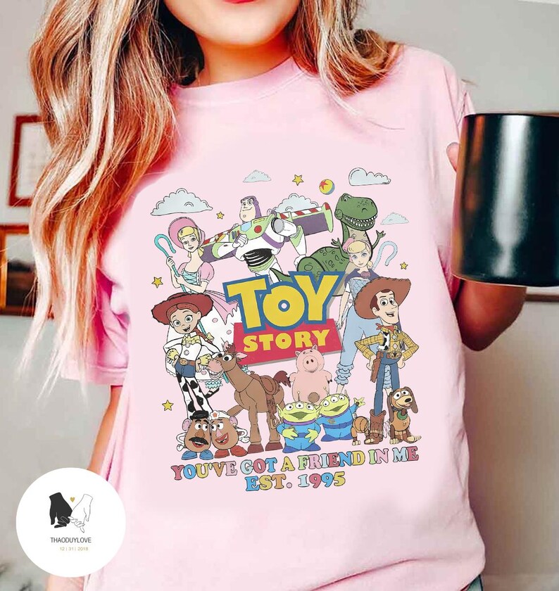 May include: Pink t-shirt with a graphic of the Toy Story characters, including Woody, Buzz Lightyear, Jessie, and Rex. The text "Toy Story" is printed in large letters, and the phrase "You've got a friend in me Est. 1995" is printed below the characters.