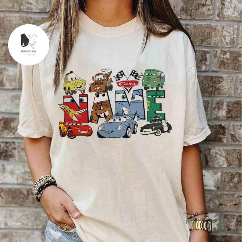 K&ouml;nnte beinhalten: Cremefarbenes T-Shirt mit dem Wort "NAME", das mit Zeichentrickfiguren von Autos geschrieben ist. Das Wort "Cars" steht &uuml;ber den Buchstaben "ME". Das Shirt hat eine lockere Passform und kurze &Auml;rmel.