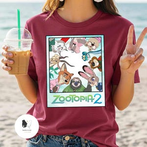 以下が含まれることがあります： アニメ映画「ズートピア2」のカラフルなグラフィックが特徴のマルーンTシャツ。デザインには様々な動物のキャラクターと、映画のタイトルがターコイズ色で描かれています。Tシャツを着た人が飲み物を持っています。