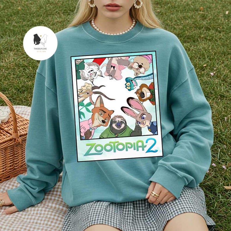 以下が含まれることがあります： アニメ映画「ズートピア2」のキャラクターが描かれた、鮮やかなグラフィックが特徴のティール色のクルーネックトレーナー。キツネ、ウサギ、その他の動物のキャラクターが含まれています。「ズートピア2」の文字は緑と白で表示されています。